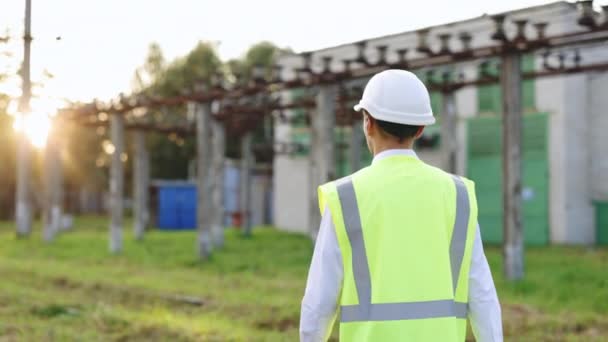 Ingegnere elettrico con casco e giubbotto di sicurezza che cammina vicino alle linee elettriche ad alta tensione verso la centrale elettrica durante il tramonto. Concetto di sviluppo di una centrale elettrica — Video Stock