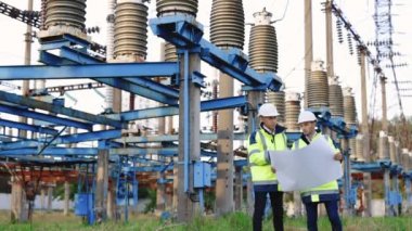 Beyaz kasklı ve yeşil yelekli iki mühendis yüksek voltajlı bir istasyonun önünde duruyorlar.