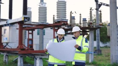 İki mühendis yüksek voltajlı elektrik hattının önündeki kağıt planına bakar, elektriğin naklini tartışır ve ellerini açık havada elektrik direklerine doğrultur.