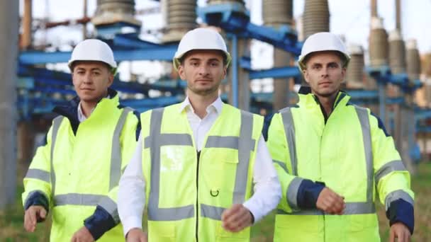 Portret grupy profesjonalnych inżynierów branży elektrycznej uśmiecha się i patrzy w kamerę. Pracownicy noszący mundur ochronny i twardy kapelusz na nieostrym tle z liniami wysokiego napięcia — Wideo stockowe