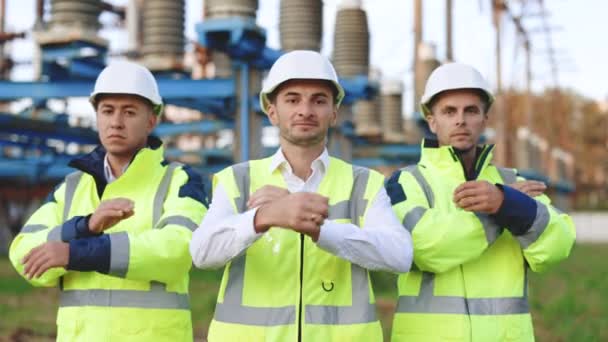 Porträtt av en grupp professionella elektriska industriingenjörer som korsar armar och tittar på kameran. Arbetstagare som bär säkerhet uniform och hatt med hög spänning elektriska ledningar bakgrund — Stockvideo
