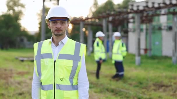 Portrét inženýra. Profesionální inženýr elektrotechniky se usmívá a dívá se do kamery. Pracovníci nosit bezpečnostní uniformu a pevný klobouk s vysokým napětím elektrických vedení pozadí — Stock video