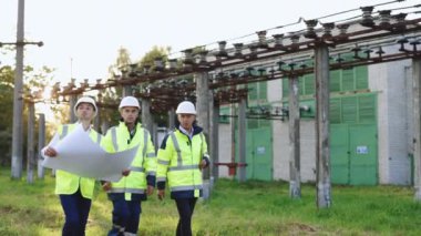 Elektrik uzmanları açık havada yeni bir proje planlıyor. Yüksek voltajlı elektrik santralinde üç mühendis elektrik hatlarının yanında yürüyor. Elektrik endüstrisi, elektrik enerjisi üretim konsepti
