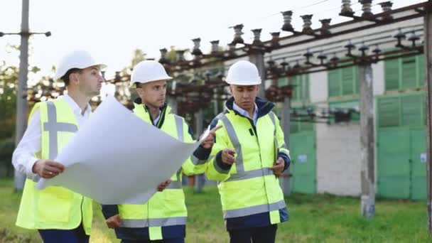 Tři inženýři procházejí v blízkosti elektráren vysokého napětí. Energetičtí specialisté plánují nový projekt venku. Elektrotechnický průmysl, koncepce výroby elektrické energie — Stock video