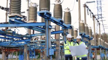 İki mühendis açık havada, yüksek voltajlı bir elektrik hattının arka planına çizilmiş bir resmi tartışıyor. Özel kıyafetli mühendisler olay yerinde çalışıyor..