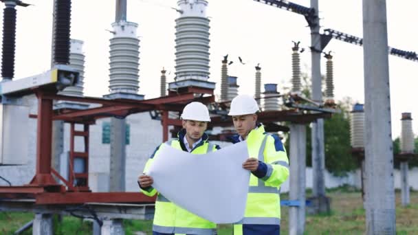 Yüksek voltajlı elektrik hattının yakınındaki mühendisler inşaat planı üzerinde çalışıyorlar. Özel kıyafetli iki mühendis, yüksek voltajlı bir elektrik hattının arka planına karşı kağıt üzerine çizim yapmayı tartışıyor. — Stok video