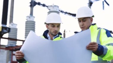 İki mühendis elektrik nakil hatlarının önünde yenilenebilir enerji geliştirme üzerinde çalışıyorlar. Özel kıyafetli mühendisler yüksek voltajlı elektrik hattının yakınındaki kağıt üzerinde bir çizimi tartıştılar.