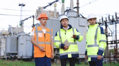 Elektrik endüstrisi, elektrik enerjisi üretim konsepti. Enerji işçileri elektrik hatlarının yakınındaki işleri tartışıyor. Bir elektrik santralinde proje planlayan özel işçiler