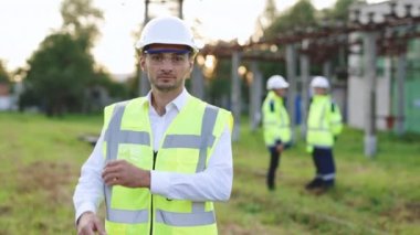Erkek mühendis teknolojisinin portresi yüksek voltajlı elektrik hatlarının arasında duruyor ve kolları göğsünün üzerinde çapraz duruyor. Koruyucu kasklı ve üniformalı bir adam gülümsüyor ve kameraya bakıyor.