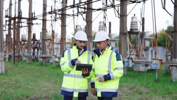 Portrait of two diverse modern factory workers wearing hardhats and uniform use digital tablet computer discussing production outdoors. High voltage station with tall pylons and voltage cables — Stock Video