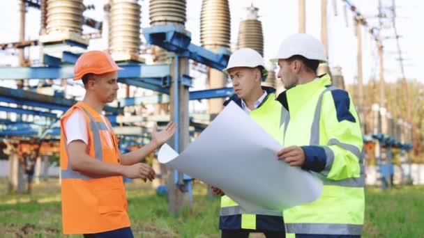 Dois Engenheiros Industriais Converse com o Trabalhador da Fábrica um desenho em papel ao ar livre contra o pano de fundo de uma usina de alta tensão. Eles trabalham na fábrica de energia elétrica — Vídeo de Stock