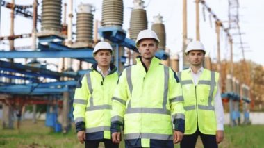 Portre mühendisleri ya da mimarlar kollarını kavuşturup kameraya bakarlar. Güvenlik üniforması giyen işçiler ve arka planda yüksek voltajlı elektrik hatları olan kasklı işçiler.