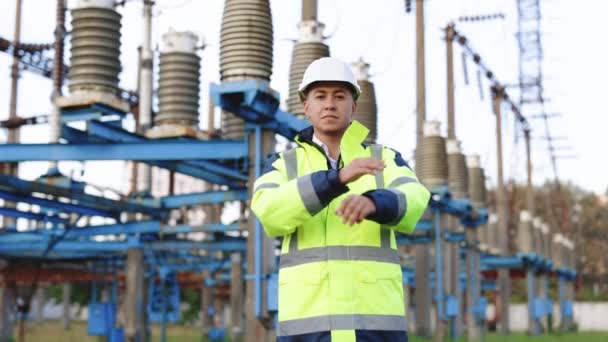 年轻微笑的亚洲电业工人的画像，手臂交叉在电源前。站在高压电站的戴硬礼帽的工程人员的画像 — 图库视频影像