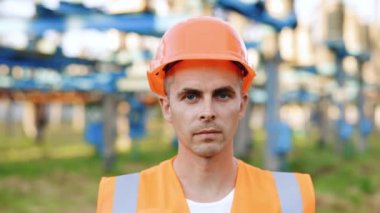 Üniformalı ve kasklı mühendisin portresi yüksek voltajlı elektrik santrali ve voltaj dağıtım kablolarının yanında duruyor.