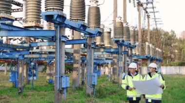 Mühendisler inşaat planını kullanırlar. Elektrik santraline karşı yürümeyi tartışırlar. Özel kıyafetli mühendisler yüksek voltajlı elektrik hattının yakınındaki kağıt üzerinde bir çizimi tartıştılar.