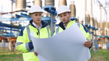 İnşaat planı olan mühendisler yüksek voltajlı elektrik hattının yanında çalışıyorlar. Özel kıyafetli iki mühendis, yüksek voltajlı bir elektrik hattının arka planına karşı kağıt üzerine çizim yapmayı tartışıyor.