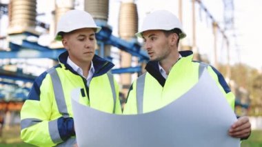 İki mühendis elektrik nakil hatlarının önünde yenilenebilir enerji geliştirme üzerinde çalışıyorlar. Özel kıyafetli mühendisler yüksek voltajlı elektrik hattının yakınındaki kağıt üzerinde bir çizimi tartıştılar.