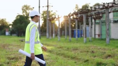 Kask ve güvenlik yeleği giyen inşaat işçisi veya mühendis elektrik santraline doğru yüksek gerilimli elektrik hatlarının yanından geçerek proje planıyla kontrol ediyor. Meslek konsepti. İş