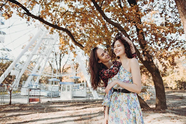 Eski moda elbiseli iki genç kadın dönme dolabın yanındaki parkta eğleniyor.