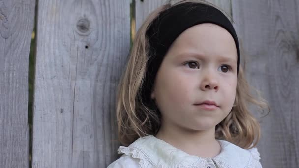 Curly-Haired Girl i en blå klänning med en svart snusnäsduk stående mot bakgrund av ett staket i trä — Stockvideo