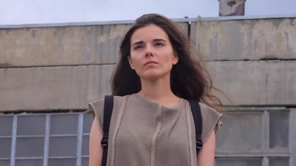Triste menina bonita em pé contra um fundo de edifícios industriais em ruínas — Vídeo de Stock