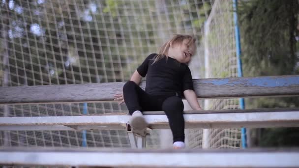 Unruhiges kleines Mädchen im schwarzen Anzug sitzt auf der Bank und springt aus dem Hintergrund Fußballgitter — Stockvideo