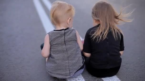 Zwei schöne fröhliche Mädchen sitzen mit dem Rücken zur Kamera auf der windigen Straße und spielen mit einem kaputten Tablet, dann schaut man sich um und steht auf und geht — Stockvideo