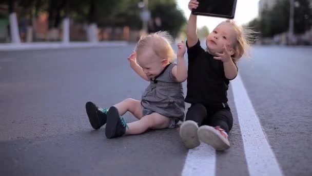 Zwei kleine positive glückliche Mädchen, die auf der Straße sitzen, das Tablet beobachten und lachen — Stockvideo