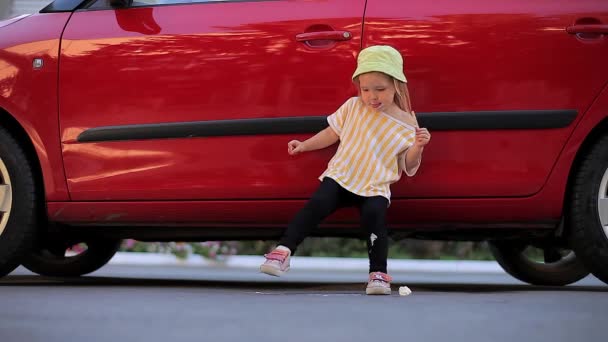 Zweijähriges lustiges Mädchen verteilt Eis auf Asphalt an den Füßen auf dem Hintergrund eines roten Autos — Stockvideo