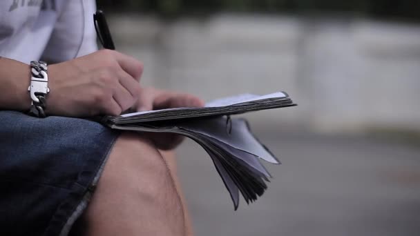 Handschrift eines Mannes in einem Notizbuch auf seinem Schoß auf der Straße — Stockvideo