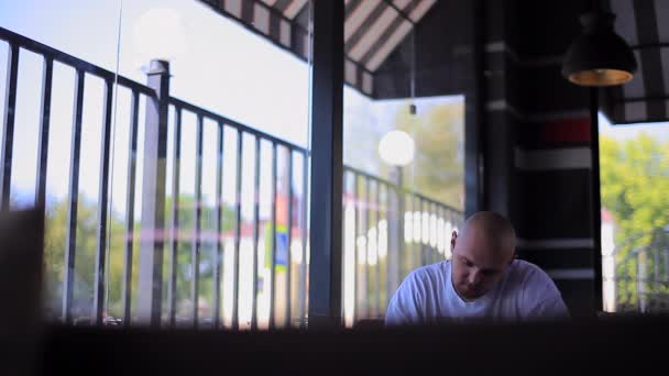 Uomo sorridente seduto in un caffè e scrive qualcosa di tanto in tanto guardando fuori dalla finestra — Video Stock