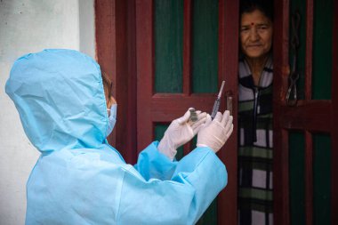 PPE koruyucu maskeli genç kadın doktor elinde şırınga ve koronavirüs aşısı tutan eldivenler, köy kadınlarıyla birlikte durup Corona virüsü aşısının önemini anlatıyor. Hastalığa karşı yeni bir aşı konsepti, grip aşısı.
