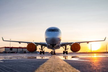 Havalimanı önlüğündeki modern yolcu uçağının ön görüntüsü resimli bir günbatımının arka planına karşıdır.