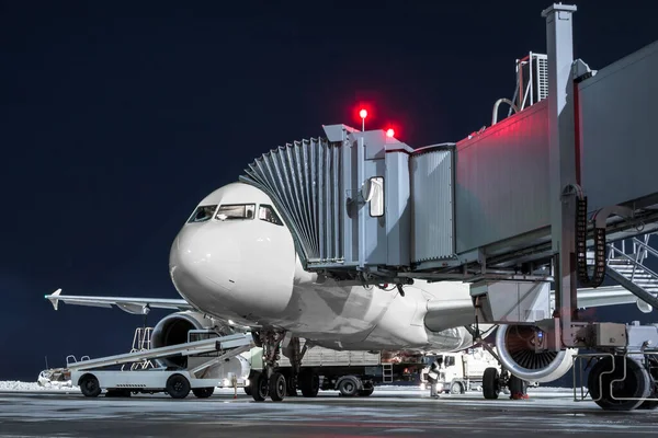Ground handling of a white passenger aircraft at the boarding bridge on the airport apron at night. Loading baggage on the airplane