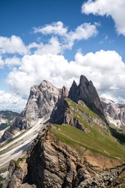Val Gardena 'daki Seceda' dan Odle tepelerinin dramatik manzarası, Dolomitler. Keskin kireçtaşı kuleleri, parlak mavi gökyüzünün altındaki yeşil alp tepelerinin üzerinde yükseliyor. İtalyan Alplerinin ikonik bir manzarası olan bulutlarla birlikte.