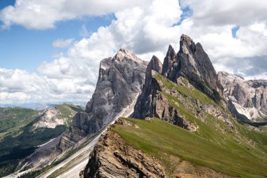Val Gardena 'daki Seceda' dan Odle tepelerinin dramatik manzarası, Dolomitler. Keskin kireçtaşı kuleleri, parlak mavi gökyüzünün altındaki yeşil alp tepelerinin üzerinde yükseliyor. İtalyan Alplerinin ikonik bir manzarası olan bulutlarla birlikte.