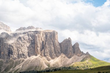Val Gardena 'daki Torri di Sella tepelerinin dramatik manzarası, Dolomitler. Keskin kireçtaşı kuleleri, parlak mavi gökyüzünün altındaki yeşil alp tepelerinin üzerinde yükseliyor. İtalyan Alplerinin ikonik bir manzarası olan bulutlarla birlikte.