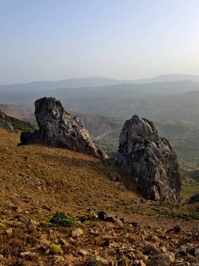 From the 1,385 m summit of Mount Ajlajel at Koudiet Ajlajel near Tetouan, Morocco, sunset spills warm light across sweeping ridgelines, rocky outcrops, and sunlit valleys and hills in the distant.