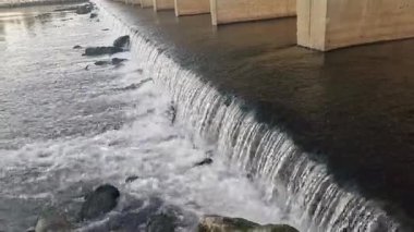 Vadideki bir köprünün altından akan geniş bir dere. Sahne suyun akışına benzer, ince yüzey detaylarını yakalar: küçük dalgalar ve girdaplar, ıslak taşların üzerinde dans eden dere.