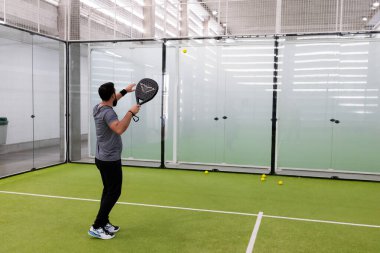 Erkek, kapalı sahada cam duvarlar ve tenis toplarıyla padel oynuyor. Yüksek kalite.