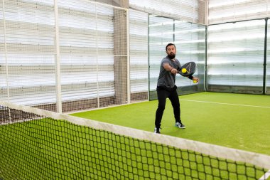 Genç İspanyol erkek kapalı sahada yüzünde odaklanmış bir ifadeyle Padel tenis oynuyor. Yüksek kalite.