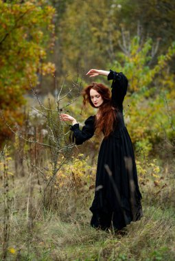 Eskimiş siyah elbiseli kızıl saçlı kadın mistik sonbahar doğası gereği uzun boylu ağaçlar, yabani dikenler ve büyüleyici düşüş atmosferi arasında zarif gotik bir şekilde poz veriyor..