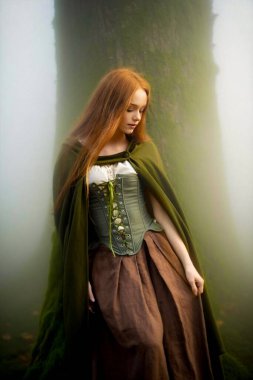 Ortaçağ yeşil pelerinli ve korseli kızıl saçlı kadın mistik yosunlu bir ormanda poz veriyor, ruhani ışık ve büyülü fantezi titreşimleriyle elf peri masalı atmosferini çağrıştırıyor..