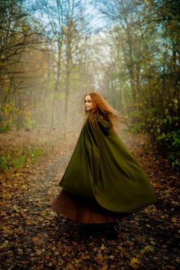 Ortaçağ yeşil pelerinli ve korseli kızıl saçlı kadın mistik yosunlu bir ormanda poz veriyor, ruhani ışık ve büyülü fantezi titreşimleriyle elf peri masalı atmosferini çağrıştırıyor..