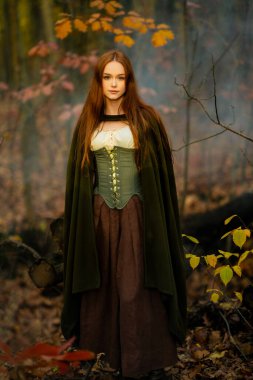 Ortaçağ yeşil pelerinli ve korseli kızıl saçlı kadın mistik yosunlu bir ormanda poz veriyor, ruhani ışık ve büyülü fantezi titreşimleriyle elf peri masalı atmosferini çağrıştırıyor..