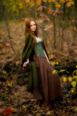 Ortaçağ yeşil pelerinli ve korseli kızıl saçlı kadın mistik yosunlu bir ormanda poz veriyor, ruhani ışık ve büyülü fantezi titreşimleriyle elf peri masalı atmosferini çağrıştırıyor..