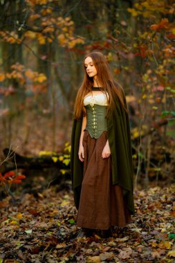 Ortaçağ yeşil pelerinli ve korseli kızıl saçlı kadın mistik yosunlu bir ormanda poz veriyor, ruhani ışık ve büyülü fantezi titreşimleriyle elf peri masalı atmosferini çağrıştırıyor..