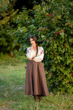 Geleneksel Ukrayna kıyafetleriyle uzun siyah örgülü güzel esmer kadın yeşil arka planda parlak kırmızı viburnum ile poz veriyor, çarpıcı bir portre, canlı bir açık hava sahnesi..