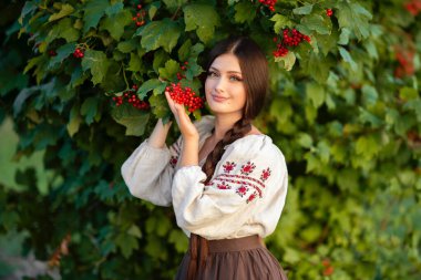 Geleneksel Ukrayna kıyafetleriyle uzun siyah örgülü güzel esmer kadın yeşil arka planda parlak kırmızı viburnum ile poz veriyor, çarpıcı bir portre, canlı bir açık hava sahnesi..
