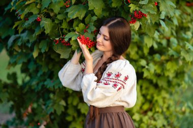 Geleneksel Ukrayna kıyafetleriyle uzun siyah örgülü güzel esmer kadın yeşil arka planda parlak kırmızı viburnum ile poz veriyor, çarpıcı bir portre, canlı bir açık hava sahnesi..